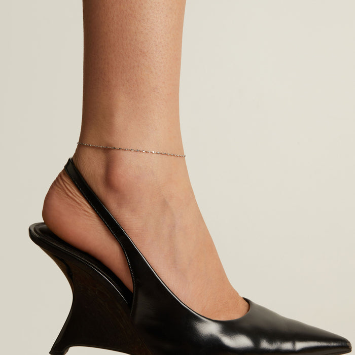 Model. Black wedge heel shoe and anklet worn by a person on a neutral background