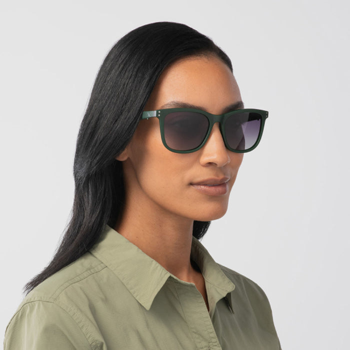 A woman with long dark hair models Eddie Bauer Velocity UV400 polarized sunglasses and a light green collared shirt, posing against a plain light background.