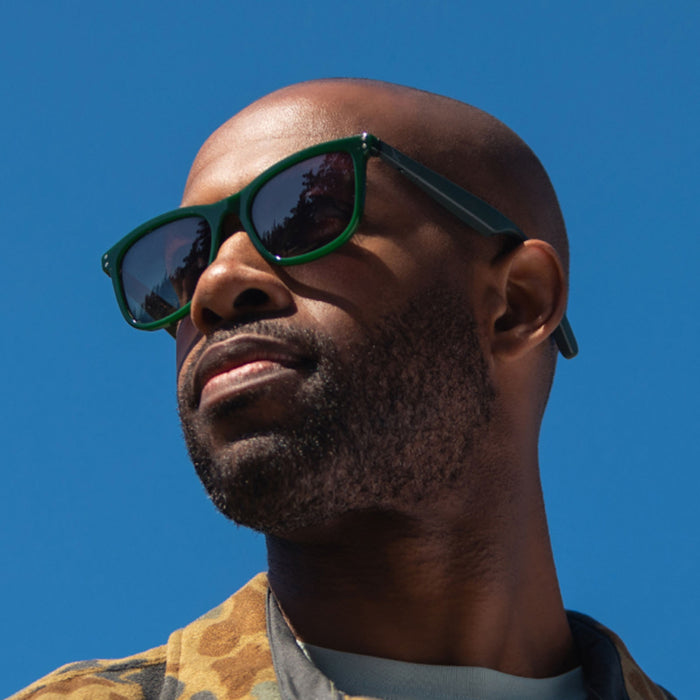 A bearded man with a shaved head wears Eddie Bauer Velocity UV400 polarized sunglasses, gazing into the distance under a clear blue sky and sporting a patterned jacket.
