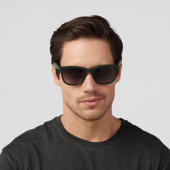 A man with short brown hair wears Eddie Bauer Velocity dark green Bluetooth audio glasses and a dark grey t-shirt, looking straight ahead against a plain light background.