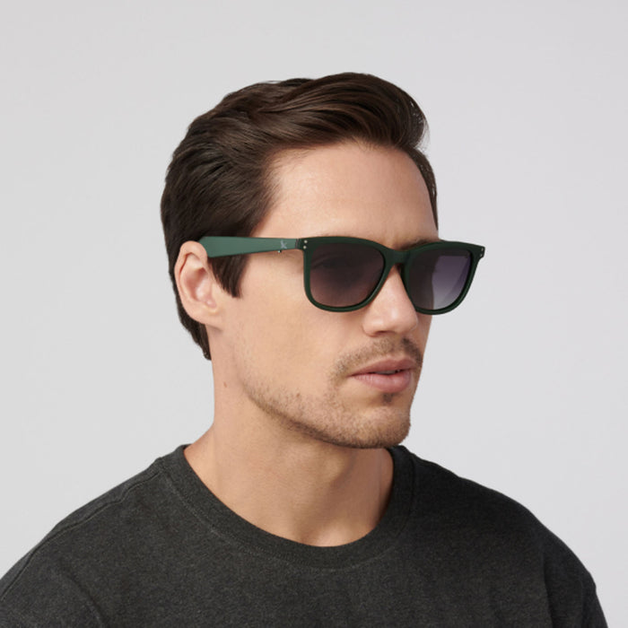 A man with short brown hair wearing Eddie Bauer Velocity Bluetooth audio glasses and a dark grey t-shirt looks slightly to the side against a light grey background.
