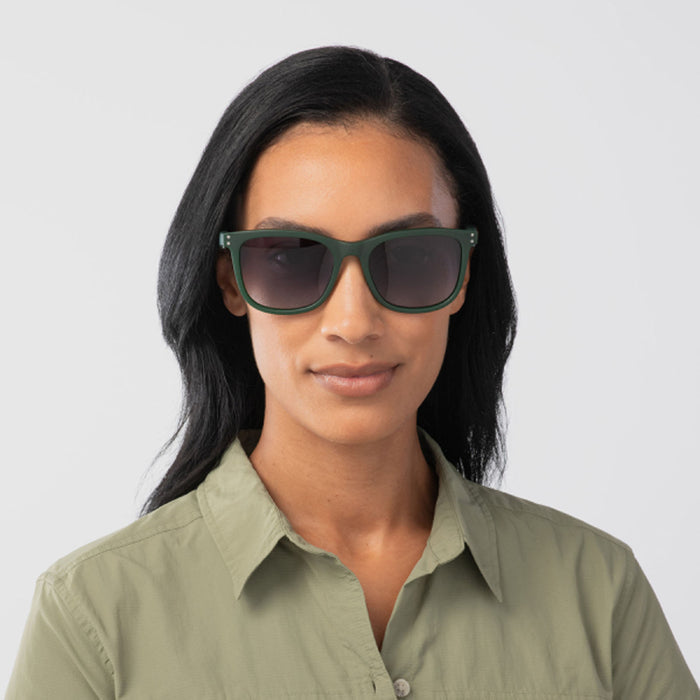A woman with long dark hair wears Eddie Bauer Velocity sunglasses with green UV400 polarized frames and a light green button-up shirt, standing against a plain light background.
