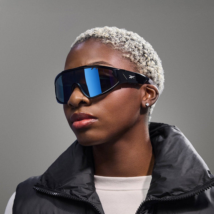 A person with short, platinum blonde hair wears Reebok Voltage sunglasses with large, reflective blue polarized lenses, small stud earrings, and a black puffer vest over a light-colored top against a plain gray background.