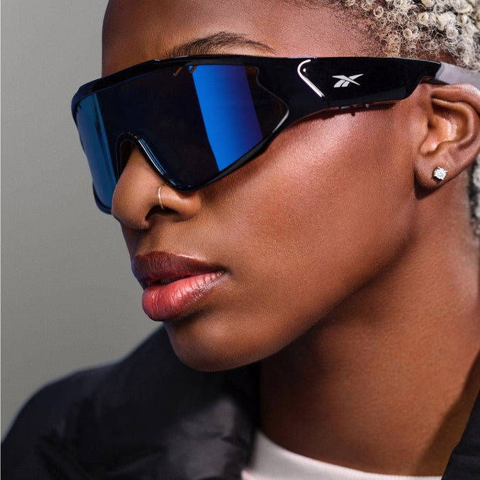 A person with light blonde curly hair wears large, reflective blue Reebok Voltage Bluetooth sport sunglasses with polarized lenses, a nose ring, stud earrings, and a black jacket, gazing confidently into the distance against a neutral background.