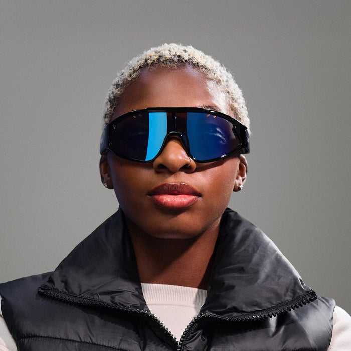 A person with short, bleached blonde hair models Reebok Voltage Bluetooth sport sunglasses with polarized lenses, stud earrings, a white top, and a black puffy vest against a plain gray background.