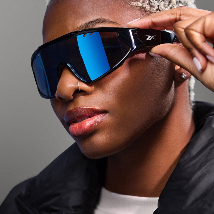 A person with short, light blonde hair wears Reebok Voltage sunglasses with large, reflective blue polarized lenses and a black jacket, holding the sunglasses at the side. The background is neutral gray.