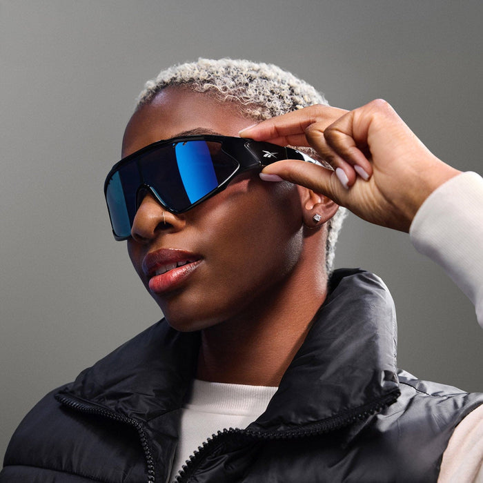 A person with short, bleached hair and a black puffer vest adjusts Reebok Voltage blue-tinted polarized lenses, gazing confidently to the side against a neutral background.