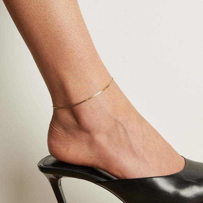 Model. Gold anklet on a foot wearing a black high heeled shoe against a neutral background.