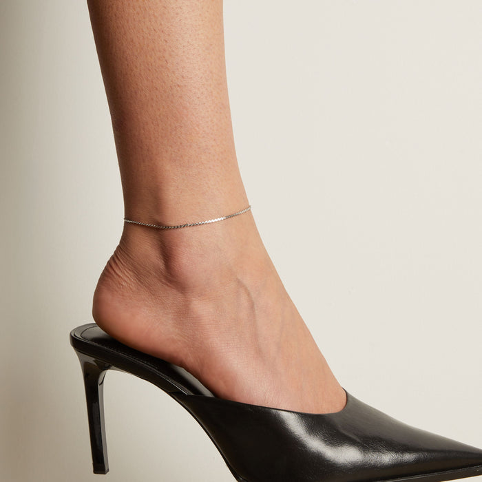 Model. Silver anklet on a foot wearing a black high heeled shoe against a neutral background.
